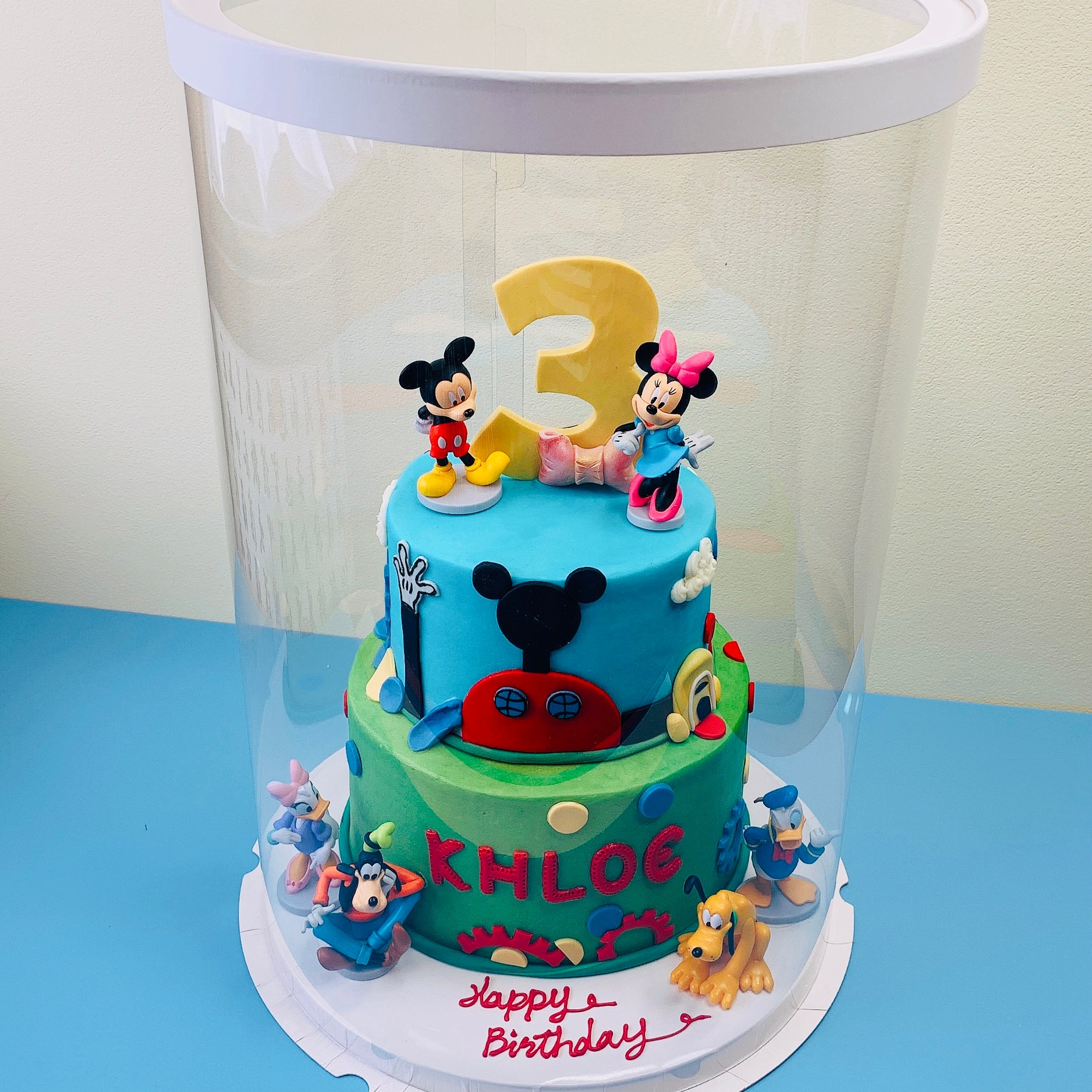 Two-tier birthday cake with cartoon characters and number 3, under a clear dome on a blue surface.