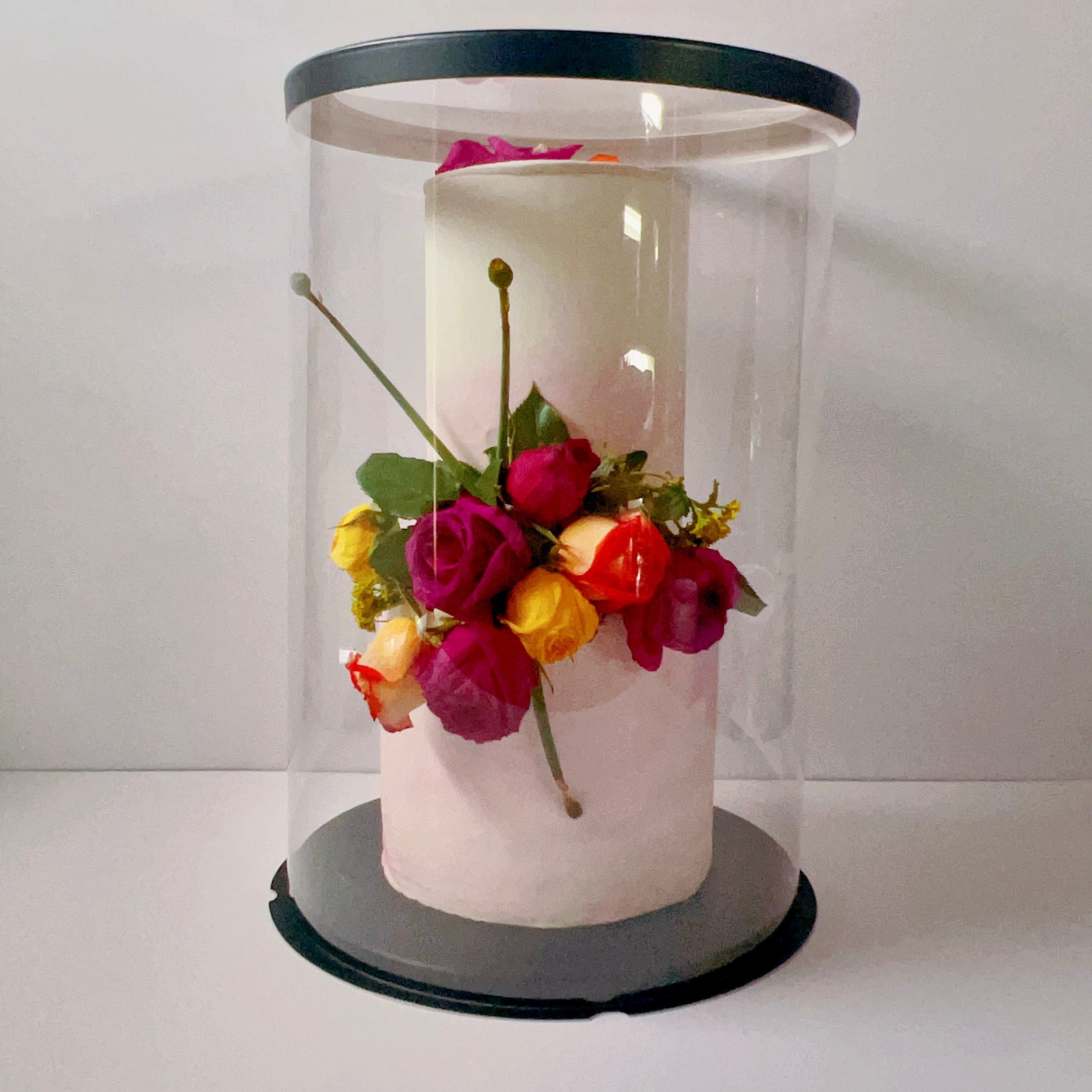 White cake with colorful flowers inside a clear cylindrical display case on a white background