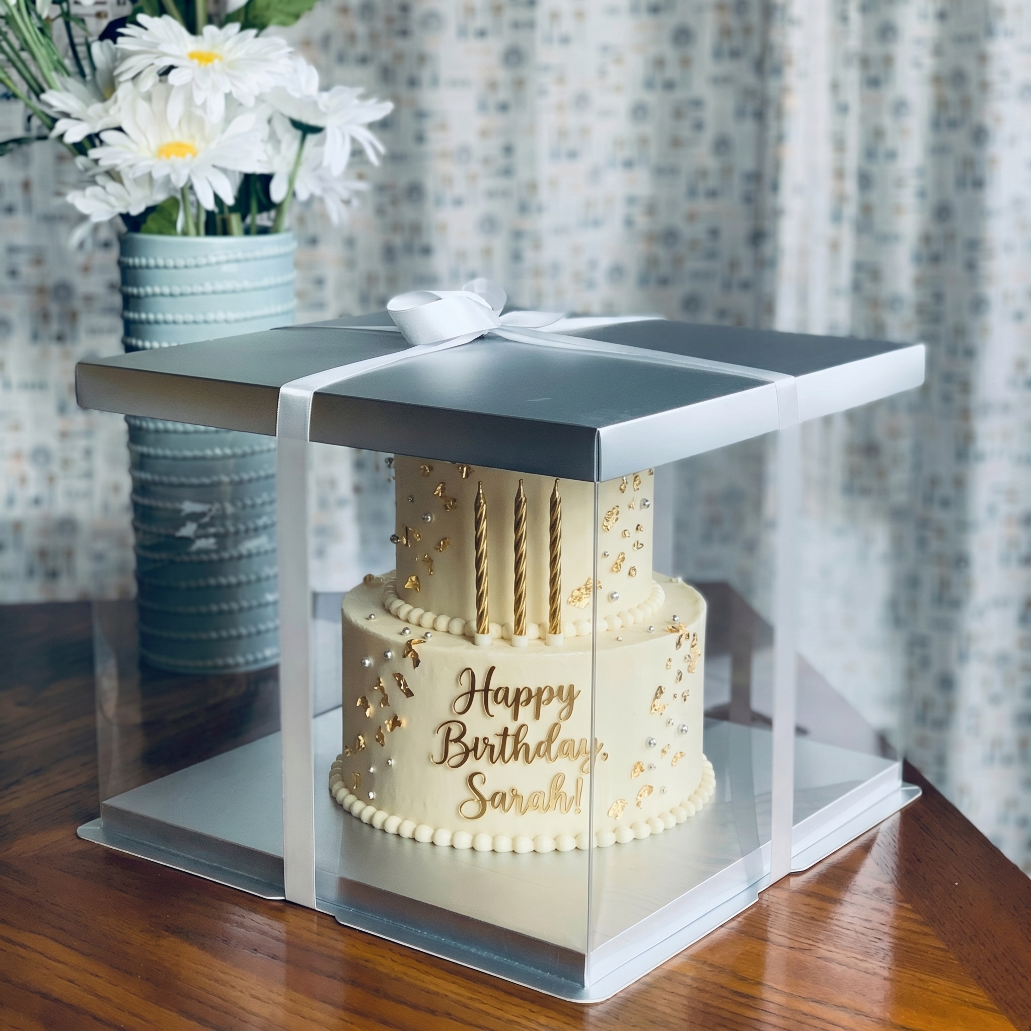 Two-tier birthday cake with 'Happy Birthday Sarah!' on a transparent cake stand, next to a vase of flowers.
