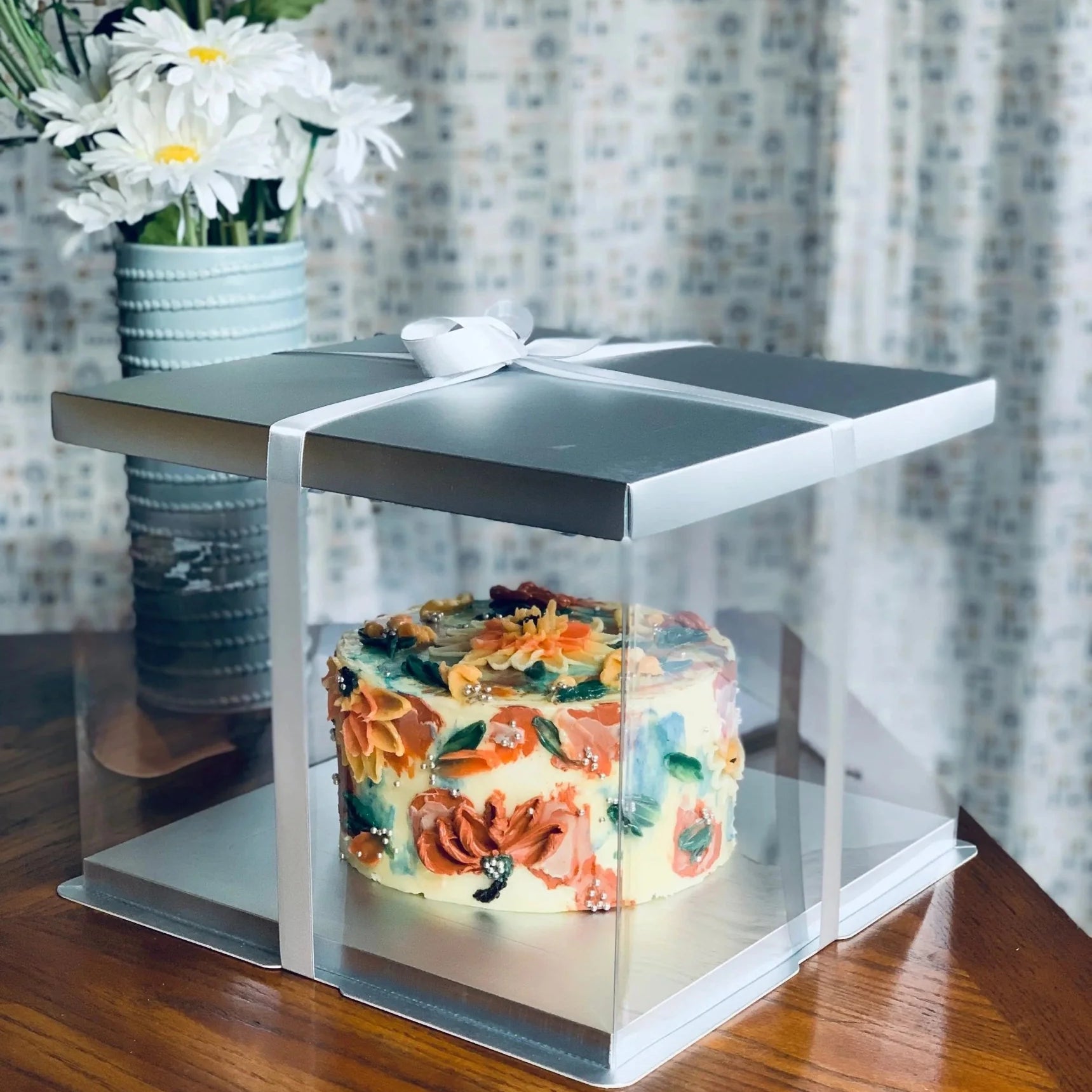 Decorative cake in a clear box with a silver lid, placed on a wooden surface with a floral arrangement in the background.