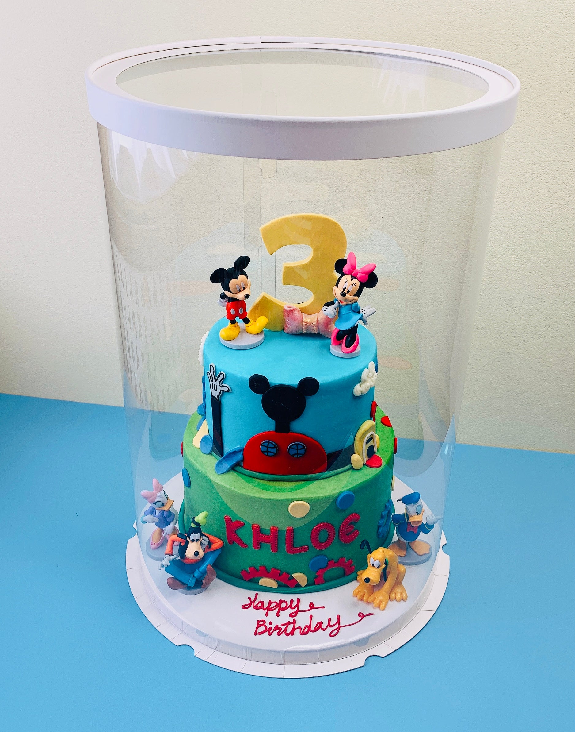 Two-tier birthday cake with cartoon characters and number 3, under a clear dome on a blue surface.