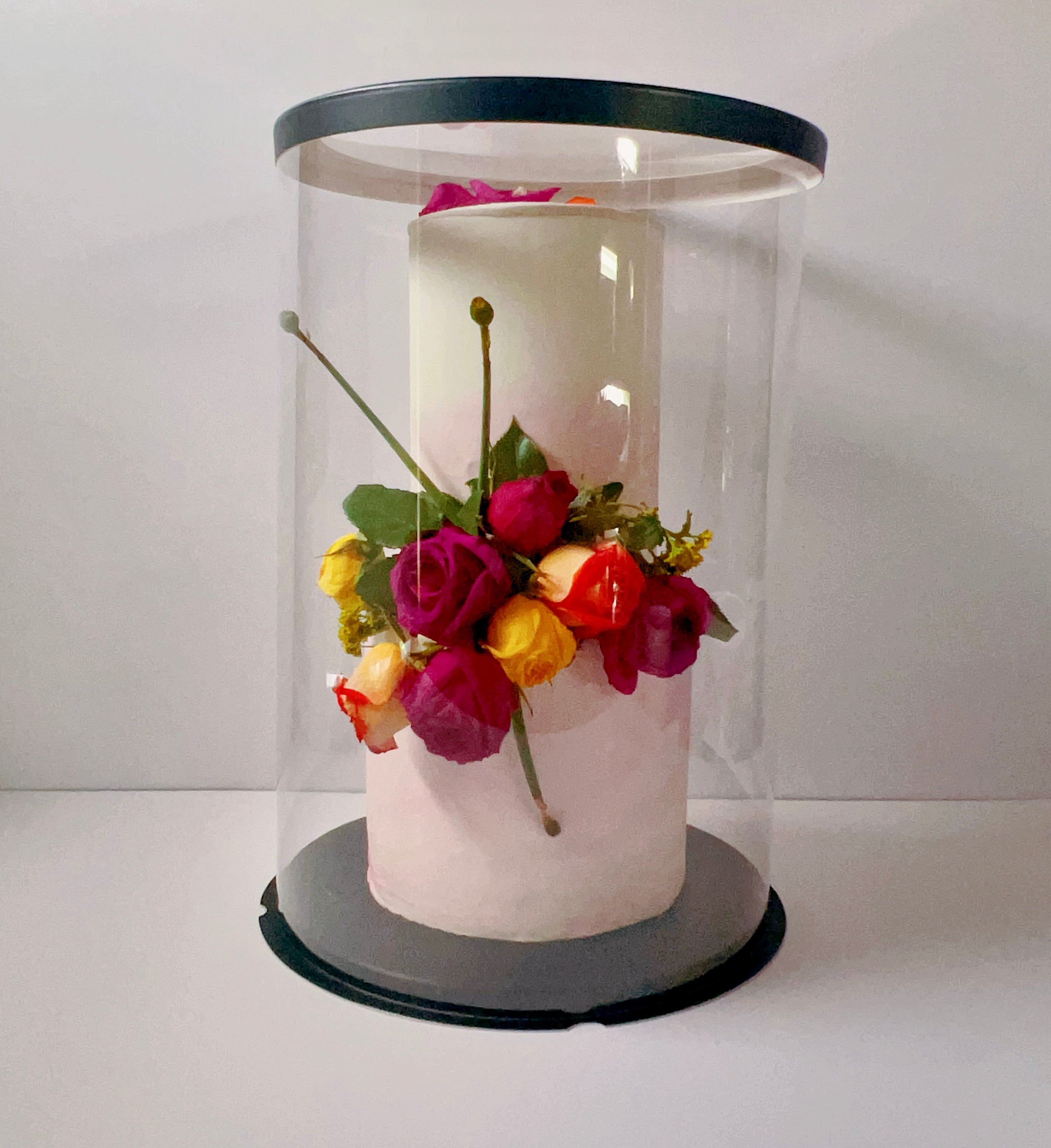 White cake with colorful flowers inside a clear cylindrical display case on a white background