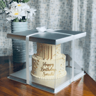 Two-tier birthday cake with 'Happy Birthday Sarah!' on a transparent cake stand, next to a vase of flowers.