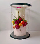 White cake with colorful flowers inside a clear cylindrical display case on a white background