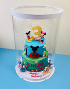 Two-tier birthday cake with cartoon characters and number 3, under a clear dome on a blue surface.
