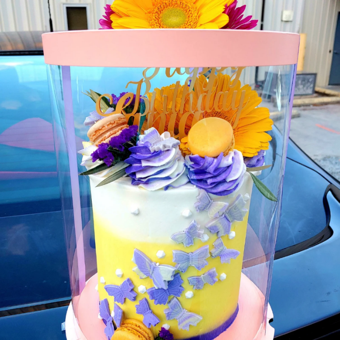 Decorative cake with flowers and butterflies in a clear container, outdoors.