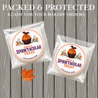 Two packaged round treats in clear cellophane bags with printed Halloween-themed labels reading 'HAVE A Spooktacular TREAT' and a cupcake illustration, shown on a gray wood plank background with large promotional text at the top.