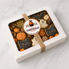 A white gift box with a clear window displaying Halloween-themed sugar cookies, including cat and ghost shapes, pumpkins, and autumn leaves, wrapped with a jute ribbon and a round 'Spooktacular Treat' label, on a light marble surface.