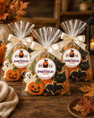Three clear cellophane bags of Halloween-themed cookies tied with cream ribbons, displayed on a rustic wooden table. The bags contain orange pumpkin-shaped cookies, black cat-shaped cookies, and white ghost-shaped cookies.