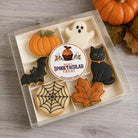 A clear plastic box of six Halloween-themed iced cookies on a gray wood surface, with a small pumpkin and fall leaves in the background. The cookies include a pumpkin, ghost, black cat, bat, spiderweb, and maple leaf shape, with a round custom label in the center reading 'Make a Spooktacular Treat'.