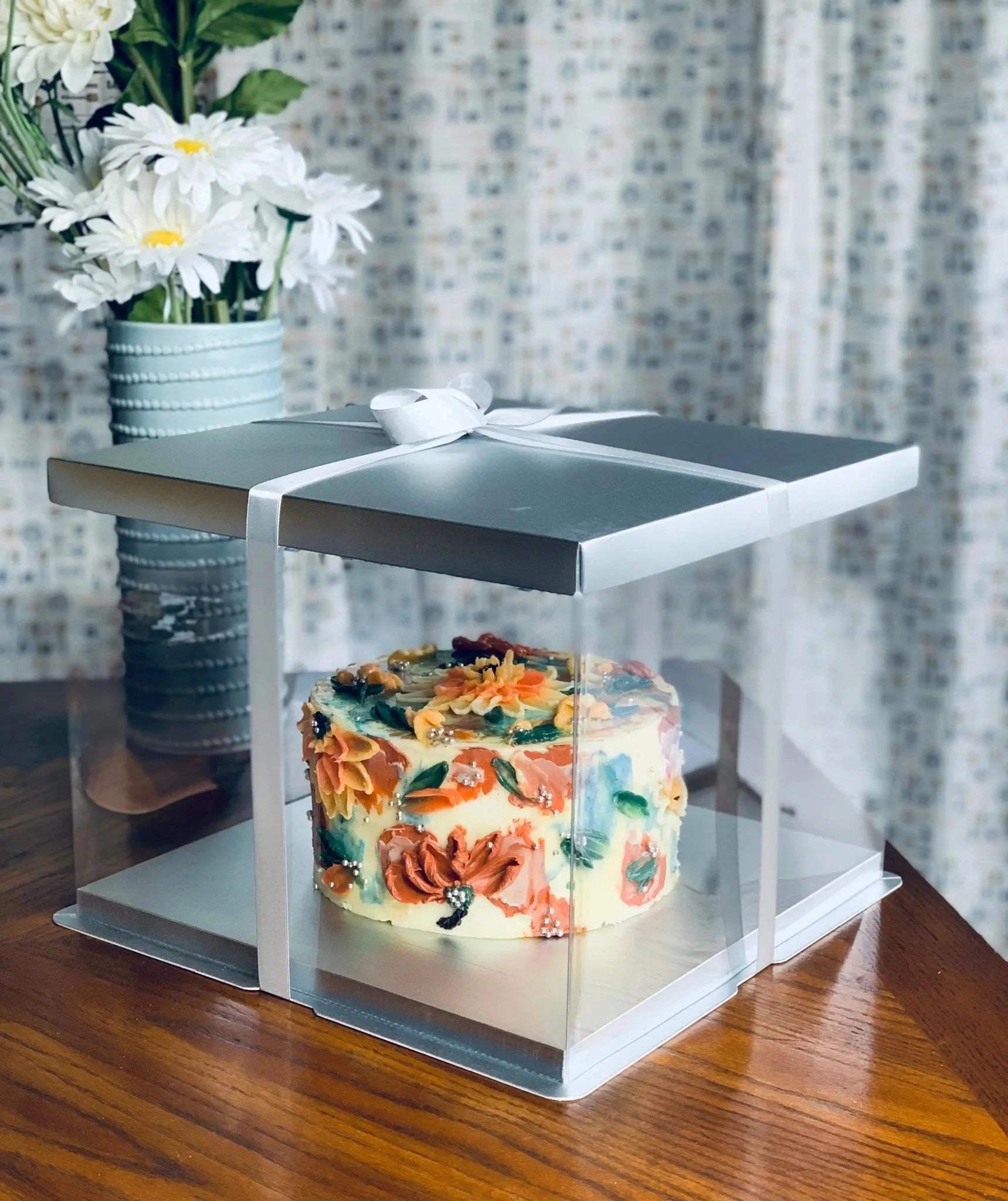 Decorative cake in a clear box with a silver lid, placed on a wooden surface with a floral arrangement in the background.