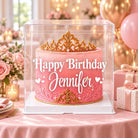 Personalized clear cake box with a pink and gold birthday cake displayed among blush balloons, roses, wrapped gifts, and party lights.