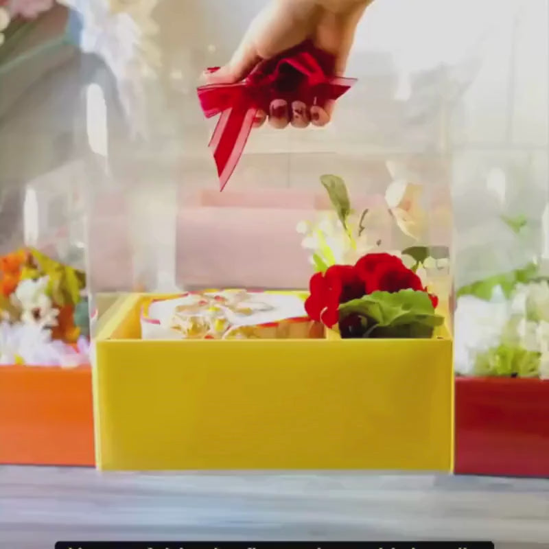 Video showing how to assemble a clear flower bento box with a yellow base, placing items inside and finishing with a red ribbon handle for presentation.
